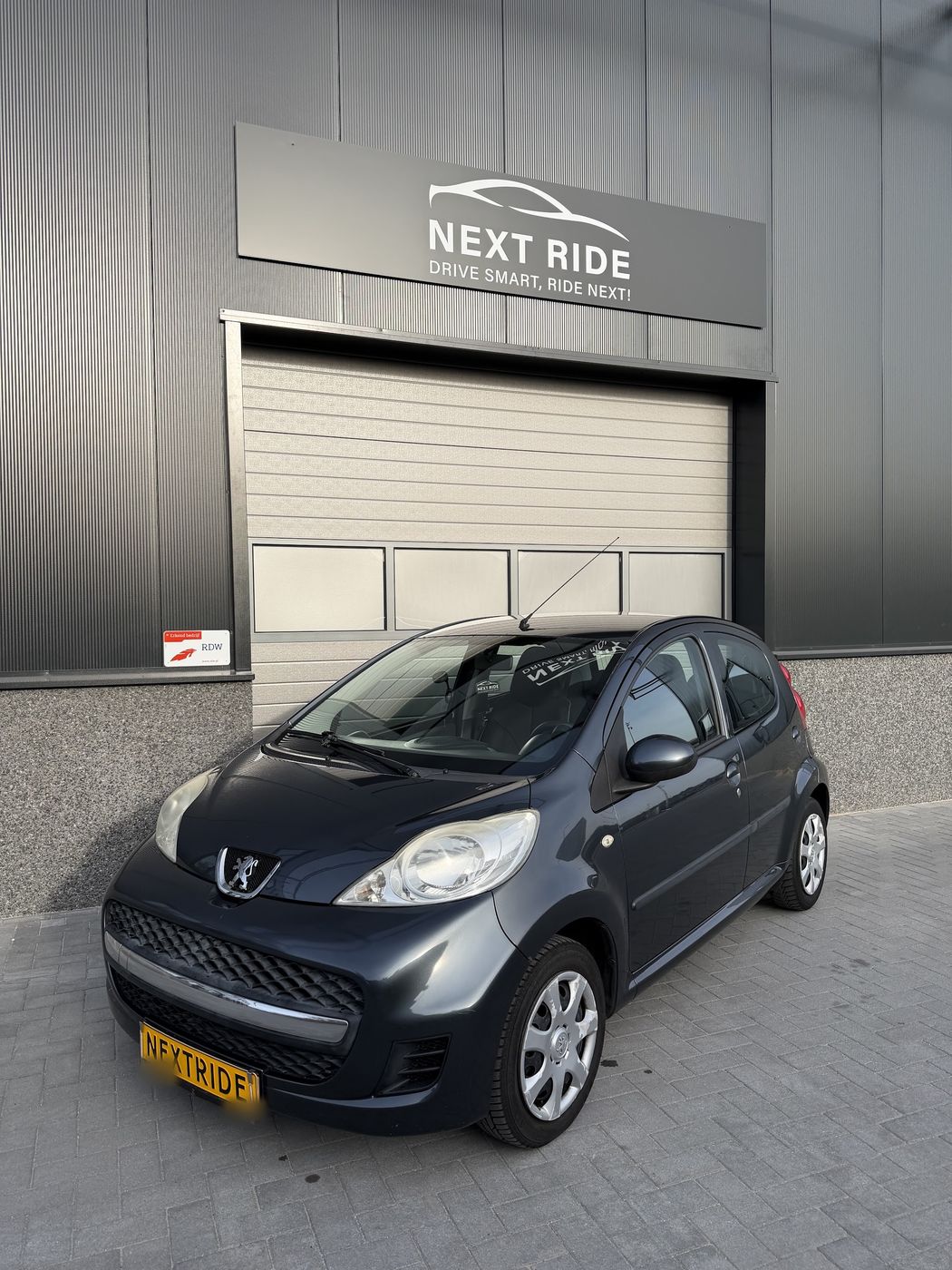 Een grijze Peugeot 107 met een geel Next Ride-bord voor de deur, naast het RDW-bordje Erkend bedrijf